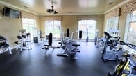 This fitness center offers a comprehensive workout environment designed to accommodate a range of exercise needs. The space is outfitted with multiple strength-training machines, including cable systems, adjustable benches, and resistance equipment, all arranged with ample room to move freely between stations. The durable rubber flooring provides both safety and comfort underfoot, making it well-suited for a variety of workout routines.

Large windows line the walls, flooding the room with natural light and offering views of the surrounding greenery, creating an open and energizing atmosphere. Patterned valance curtains add a touch of style to the otherwise functional space. A decorative ceiling fan is centrally mounted, ensuring steady airflow and maintaining a comfortable temperature during workouts.

The warm, neutral-toned walls and crown molding give the fitness center a refined aesthetic that sets it apart from a typical gym environment. The overall layout is thoughtfully organized, allowing multiple users to exercise simultaneously without feeling cramped. Whether focused on strength training or cardiovascular conditioning, this facility provides the essential equipment and a welcoming ambiance to support an active lifestyle.