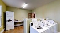 This shared laundry facility offers a practical and well-organized space designed to meet everyday laundering needs. The room is finished with soft sage-green walls and durable tile flooring, creating a clean and functional environment. A row of top-load washing machines lines one side of the room, accompanied by stacked dryer units positioned along the adjacent wall, providing ample capacity for multiple users. A folding table offers a convenient surface for sorting and folding laundry, keeping the workflow efficient. Overhead fluorescent lighting ensures the space is well-lit throughout. A windowed door with warm wood trim leads to an adjoining area, allowing for easy access and natural light to filter in. The layout is thoughtfully arranged to maximize usability while maintaining an open, uncluttered feel. Overall, this laundry room is a straightforward, no-frills facility built for convenience and practicality.