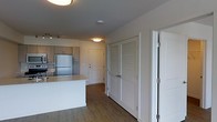 This single-floor unit is designed with a practical, open-concept layout that integrates the kitchen and living areas into one cohesive space. The kitchen is outfitted with warm-toned wood cabinetry, stainless steel refrigerator, and a sleek white island that provides additional counter space and casual seating potential. Track lighting above the kitchen adds focused illumination to the workspace, while flush-mount ceiling fixtures illuminate the adjoining hallway area.

Wood-style plank flooring runs throughout the space, adding warmth and visual continuity. The hallway connecting the main living area to the rest of the unit features a set of louvered double doors, likely concealing a storage closet or laundry area, alongside a solid paneled door providing access to an additional room. White trim and neutral wall tones create a clean, bright aesthetic throughout.

The unit's layout emphasizes functionality, with clearly defined zones for cooking, storage, and circulation. The overall ambiance is modern and understated, well-suited for everyday living.