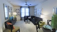 This single-floor residential space offers a thoughtfully designed and inviting interior that blends comfort with contemporary style. The living area serves as the centerpiece of the home, showcasing plush carpeted flooring and neutral-toned walls that create a warm, welcoming ambiance. A ceiling fan with a light fixture is mounted overhead, providing both air circulation and subtle illumination to complement the natural light that streams in through large, blinded windows dressed with elegant draping curtains.

The living room is furnished with a spacious navy blue sectional sofa adorned with decorative throw pillows, anchored by a pair of metallic accent tables that add a chic, modern touch. Colorful botanical and floral wall art panels flank the windows, infusing the space with personality and visual interest. Additional seating and accent furniture — including a dark dining chair and coordinating side tables with table lamps — contribute to a cohesive and layered design aesthetic.

Crown molding along the upper walls adds an architectural detail that elevates the overall finish of the space. The layout feels open and well-proportioned, allowing for easy movement and flexible furniture arrangement. Overall, this space presents a clean, stylish, and functional living environment suited for everyday comfort.