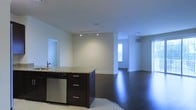 This single-floor space is designed with an open-concept layout that seamlessly connects the kitchen and living areas, creating a bright and airy atmosphere throughout. The kitchen is anchored by a central island featuring a granite countertop with warm tonal variations, complemented by rich dark wood cabinetry equipped with brushed nickel hardware. A stainless steel dishwasher and sink add a contemporary, functional touch to the space. Track lighting overhead provides focused illumination, while recessed ceiling lights contribute to the overall brightness of the interior. The transition from the tiled kitchen area to the expansive dark hardwood floors of the living space adds a striking visual contrast that enhances the modern aesthetic. The living area is generous in scale, with clean white walls and high ceilings that amplify the sense of openness. A window brings in natural light, adding warmth to the otherwise crisp, neutral palette. Neutral-toned walls throughout give the space a clean and versatile backdrop, suitable for a variety of interior styles. The overall ambiance is sleek, modern, and welcoming, with thoughtful design details that balance both form and function across the entire floor plan.