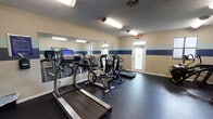 This dedicated fitness room is designed to support a comprehensive workout routine, offering a well-organized and functional space for exercise. The room features durable rubber flooring throughout, providing a safe and supportive surface for all types of physical activity. The walls are finished in a neutral beige tone accented by bold navy and light blue horizontal stripes, giving the space a clean and energetic aesthetic. Fluorescent lighting fixtures illuminate the room evenly, complemented by natural light from several windows. A large wall mirror on one side of the room enhances the sense of space while allowing users to monitor their form during workouts. Cardio equipment includes treadmills, a stationary bike, an elliptical, and a stair climber, while strength training needs are met with a multi-station cable machine, a rack of dumbbells in varying weights, and an assortment of medicine balls and resistance accessories. A wall-mounted flat-screen TV adds an entertainment element to the space. The room is accessed through a frosted-glass entry door and features a textured ceiling with ample ventilation. Overall, the fitness room provides a well-rounded environment suitable for a full range of exercise disciplines.