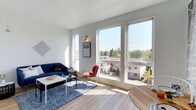 This single-floor space showcases a clean, contemporary design defined by an abundance of natural light and an open, airy atmosphere. The living area is the focal point of the home, anchored by a large bank of floor-to-ceiling windows that frame expansive views of the surrounding neighborhood rooftops and a lush tree canopy beyond. These oversized windows flood the interior with sunlight throughout the day, creating a bright and welcoming environment. A secondary narrow vertical window on the adjacent wall adds an additional source of natural light, enhancing the sense of openness.

The interior features light hardwood flooring that runs throughout, complementing the crisp white walls and contributing to the space's modern aesthetic. The living area comfortably accommodates a sofa, accent chair, coffee table, and area rug, demonstrating a well-proportioned layout. A dining area is partially visible, furnished with a round wooden table. Wall-mounted accent lighting adds a warm, functional touch. The overall ambiance is bright, minimalist, and contemporary, with the outdoor views serving as a standout architectural feature of the space.