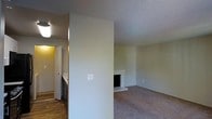 This single-floor residence offers a straightforward and functional layout designed for comfortable everyday living. The living area is finished with wall-to-wall carpeting, providing a warm and inviting atmosphere, and is anchored by a classic fireplace with a light-colored surround that serves as a natural focal point of the room. A partial dividing wall separates the living space from the adjacent kitchen area, creating a sense of definition between the two zones while maintaining an open feel. The kitchen features hardwood-style flooring, adding a contrasting texture to the softer carpeted living space, and includes cabinetry and standard appliances. Louvered bifold doors are visible near the kitchen, likely concealing a utility or storage area. Throughout the space, a textured popcorn ceiling adds a consistent overhead detail. Neutral, light-toned walls in both areas contribute to a clean and airy aesthetic, offering a versatile canvas for personalization. The overall layout is compact yet well-organized, making efficient use of the available space across the single floor.