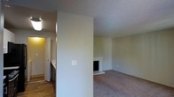 This single-floor residence offers a straightforward and functional layout designed for comfortable everyday living. The living area is finished with wall-to-wall carpeting, providing a warm and inviting atmosphere, and is anchored by a classic fireplace with a light-colored surround that serves as a natural focal point of the room. A partial dividing wall separates the living space from the adjacent kitchen area, creating a sense of definition between the two zones while maintaining an open feel. The kitchen features hardwood-style flooring, adding a contrasting texture to the softer carpeted living space, and includes cabinetry and standard appliances. Louvered bifold doors are visible near the kitchen, likely concealing a utility or storage area. Throughout the space, a textured popcorn ceiling adds a consistent overhead detail. Neutral, light-toned walls in both areas contribute to a clean and airy aesthetic, offering a versatile canvas for personalization. The overall layout is compact yet well-organized, making efficient use of the available space across the single floor.