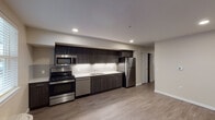 Located at 2220 SW 192nd Ave in Vancouver, Washington, this single-floor unit offers a clean, modern aesthetic throughout. The kitchen serves as a standout feature, equipped with a full suite of stainless steel appliances including a gas range, built-in microwave, dishwasher, and refrigerator. Rich dark cabinetry extends to the ceiling, providing generous storage, while crisp white countertops and a classic subway tile backsplash offer a contemporary contrast. Recessed lighting illuminates the space efficiently throughout. The open layout connects the kitchen seamlessly to the living area, which is finished with durable wood-look flooring that carries throughout the unit. A dedicated living space features neutral walls, recessed lighting, and a wall-mounted thermostat, offering a blank canvas for personalization. Natural light filters in through double-pane windows, with a wall-mounted HVAC unit ensuring year-round comfort. The bathroom is well-appointed with a full tub and shower combination, a vanity with a white countertop, and clean neutral finishes. The overall design emphasizes functionality and modern simplicity, with consistent flooring, neutral tones, and quality finishes tying each room together cohesively.