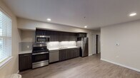 Located at 2220 SW 192nd Ave in Vancouver, Washington, this single-floor unit offers a clean, modern aesthetic throughout. The kitchen serves as a standout feature, equipped with a full suite of stainless steel appliances including a gas range, built-in microwave, dishwasher, and refrigerator. Rich dark cabinetry extends to the ceiling, providing generous storage, while crisp white countertops and a classic subway tile backsplash offer a contemporary contrast. Recessed lighting illuminates the space efficiently throughout. The open layout connects the kitchen seamlessly to the living area, which is finished with durable wood-look flooring that carries throughout the unit. A dedicated living space features neutral walls, recessed lighting, and a wall-mounted thermostat, offering a blank canvas for personalization. Natural light filters in through double-pane windows, with a wall-mounted HVAC unit ensuring year-round comfort. The bathroom is well-appointed with a full tub and shower combination, a vanity with a white countertop, and clean neutral finishes. The overall design emphasizes functionality and modern simplicity, with consistent flooring, neutral tones, and quality finishes tying each room together cohesively.