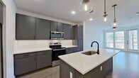 This well-designed one-bedroom, one-bathroom unit offers a thoughtful layout across a single floor, making efficient use of its space. The open-concept kitchen and living area serves as the heart of the unit, featuring light wood-tone flooring, recessed lighting, and a ceiling fan. The kitchen is a standout, equipped with charcoal gray flat-front cabinetry extending to the ceiling for generous storage, white quartz countertops, a subway tile backsplash, and a full suite of stainless steel appliances including a French door refrigerator, electric range, and built-in microwave. A central island with an undermount sink and matte black faucet provides additional prep space and pendant lighting overhead adds a modern touch. The living area opens to a balcony through large windows and a glass door, allowing abundant natural light and outdoor views. The bedroom features plush carpeting, a ceiling fan, and a window with exterior views. The bathroom offers a white quartz vanity with dark cabinetry, a walk-in glass-enclosed shower with marble-style tile surround, and light stone-look tile flooring throughout, completing the modern aesthetic.