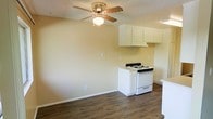 This single-floor space offers a practical and straightforward layout that blends the living area and kitchen into one cohesive, open environment. The dark hardwood-style flooring runs throughout, providing a warm contrast to the light, neutral-toned walls that lend the space a clean and airy feel.

The kitchen is equipped with a white gas range and complemented by white cabinetry overhead, offering functional storage. An under-cabinet range hood is integrated neatly into the design, keeping the kitchen area tidy and efficient.

The living area benefits from a ceiling fan with a built-in light fixture, which promotes air circulation and adds ambient lighting. The textured ceiling adds a layer of classic character to the space. Multiple electrical outlets are conveniently positioned along the walls, adding to the overall functionality.

The open-concept nature of the layout maximizes the sense of space, making the interior feel welcoming and easy to navigate. The neutral color palette throughout creates a versatile backdrop, well-suited to a variety of interior styles. Overall, this space delivers a simple, functional living environment with cohesive design elements throughout.