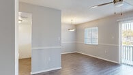This single-floor residence offers a clean, neutral aesthetic throughout, with light gray walls accented by white chair-rail molding that runs consistently across the main living areas. The open living and dining space is well-lit, drawing in natural light through a large sliding glass door fitted with vertical blinds that opens to an outdoor view, complemented by a window with shuttered blinds. A ceiling fan with a built-in light fixture serves the living area, while a pendant light defines the dedicated dining space nearby. Wood-look plank flooring extends throughout the living, dining, and kitchen areas, lending a cohesive, easy-to-maintain surface underfoot. The galley-style kitchen features white cabinetry, light countertops, a full suite of white appliances including a range, dishwasher, and refrigerator, along with a under-cabinet range hood and a tiled backsplash. The bedroom is carpeted for comfort and includes two side-by-side closets with shelving and hanging rods, providing generous storage. The bathroom presents a tub-shower combination with tile surround, a white vanity with storage below, and wood-look flooring, maintaining the unit's bright, clean character throughout.