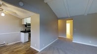 This single-floor space is defined by its striking vaulted ceiling with exposed wooden beams, which lend architectural character and create a sense of generous vertical volume in the main living area. The open layout seamlessly connects the living space to the kitchen, which is outfitted with white cabinetry, dark countertops, and stainless steel appliances, including a range and over-the-range microwave. The kitchen features hardwood flooring, while the living area transitions to plush carpeting, adding warmth and comfort underfoot. Neutral gray walls run throughout, providing a clean, versatile backdrop. A hallway visible from the main living area leads to additional rooms, with natural light visible at the end suggesting well-lit secondary spaces. White baseboards and trim provide crisp contrast against the gray walls, contributing to a tidy, cohesive aesthetic. The overall ambiance is bright, airy, and functional, with the vaulted ceiling serving as the standout architectural feature that elevates the character of the entire space.
