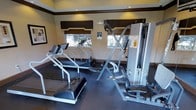 This fitness room is designed to support a well-rounded workout routine within a efficiently organized single-level space. The room features a dark, rubberized flooring ideal for heavy equipment use, providing both durability and safety during exercise. Two treadmills are positioned toward the windowed wall, allowing users to enjoy natural light and outdoor views while engaging in cardio sessions. A multi-function cable strength training machine occupies a central position in the room, offering a variety of resistance-based exercises. Two windows dressed with structured Roman shades allow ample natural light to filter in, creating a bright and welcoming atmosphere. Recessed ceiling lights supplement the natural light, ensuring the space remains well-lit at all hours. The warm-toned walls contribute to an inviting environment, while the dark wood window trim and valances add a polished, finished aesthetic. The room is compact yet functional, with equipment thoughtfully arranged to maximize usable floor space. Framed wall art adds a subtle decorative touch, balancing the utilitarian nature of the fitness equipment with a more refined ambiance.