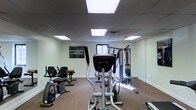 This dedicated fitness space is designed to support a comprehensive workout experience within a single-floor layout. The room features a drop ceiling with large recessed panel lighting that provides bright, even illumination throughout, creating a clean and functional environment. The flooring is finished with a durable, speckled carpet suitable for high-traffic exercise use.

The space is outfitted with a variety of exercise equipment, including a stationary recumbent bike, an elliptical cross-trainer, and a multi-station cable and weight machine along the far wall, enabling both cardiovascular and strength training activities. A window along the back wall allows for a degree of natural light to complement the overhead fixtures.

A motivational wall banner reading "The only bad workout is the one that didn't happen" adds an energizing touch to the atmosphere. Additional accessories and resistance training aids are visible near the multi-station unit, rounding out the room's offerings. The walls are painted a neutral off-white, keeping the space feeling open and uncluttered. Overall, this fitness room is thoughtfully arranged to maximize usable floor space while accommodating a diverse range of exercise equipment for a well-rounded training environment.