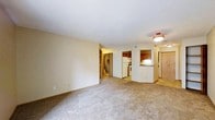This single-floor residence presents an open and functional layout visible from the spacious carpeted living area. The living space features neutral beige walls, light-colored carpet flooring, and standard-height ceilings with flush-mount lighting, creating a clean and understated ambiance. The open sightlines from the living area extend toward the kitchen, which is accessible via a half-wall pass-through that adds a sense of connectivity between the two spaces. The kitchen is equipped with dark wood cabinetry, a white refrigerator, and a range, providing practical functionality. Countertop space is available along the kitchen's perimeter, with under-cabinet lighting adding subtle warmth to the area. A hallway branching off from the main living space leads to additional rooms, including what appears to be a closet or utility area with bifold doors and a stairwell visible in the background. The entryway, partially visible to the right, features a light wood-toned door and tile flooring, offering a transitional space into the unit. Overall, the space maintains a simple, neutral palette throughout, making it a versatile and move-in ready environment.