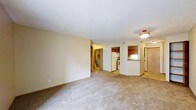 This single-floor residence presents an open and functional layout visible from the spacious carpeted living area. The living space features neutral beige walls, light-colored carpet flooring, and standard-height ceilings with flush-mount lighting, creating a clean and understated ambiance. The open sightlines from the living area extend toward the kitchen, which is accessible via a half-wall pass-through that adds a sense of connectivity between the two spaces. The kitchen is equipped with dark wood cabinetry, a white refrigerator, and a range, providing practical functionality. Countertop space is available along the kitchen's perimeter, with under-cabinet lighting adding subtle warmth to the area. A hallway branching off from the main living space leads to additional rooms, including what appears to be a closet or utility area with bifold doors and a stairwell visible in the background. The entryway, partially visible to the right, features a light wood-toned door and tile flooring, offering a transitional space into the unit. Overall, the space maintains a simple, neutral palette throughout, making it a versatile and move-in ready environment.