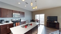 This single-floor residence showcases an open-concept layout that seamlessly connects the kitchen, dining area, and living space. The kitchen is equipped with dark espresso cabinetry, stainless steel appliances including a range and over-the-range microwave, and a generous island topped with light quartz countertops featuring subtle veining. A deep undermount sink with a modern arc faucet anchors the island, which provides both prep space and casual seating potential. Pendant lighting with cylindrical frosted shades hangs above the island, complementing the recessed lighting throughout the ceiling. The flooring features dark hardwood-style planks that contrast elegantly with the light gray and taupe wall tones. The dining area accommodates a full table and chairs, positioned directly adjacent to a sliding glass door that opens to a balcony, flooding the space with natural light and offering outdoor views. The open sightlines between the kitchen, dining, and living areas create a cohesive, functional environment well-suited for both everyday living and entertaining.