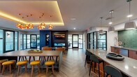 This thoughtfully designed single-floor amenity space blends modern aesthetics with functional communal living. Throughout, a striking herringbone-patterned wood-look floor creates a cohesive visual foundation, complemented by a consistent palette of warm woods, dark accents, and neutral tones.

The main lounge and social area serves as the heart of the space, featuring a dramatic coffered ceiling with warm ambient uplighting and a cluster of globe pendant lights in brushed gold. A dark-paneled fireplace wall anchors the room, paired with a wall-mounted display screen and sculptural wall art. Multiple seating configurations — including leather barrel chairs, bar-height seating along a large island, and low stools at communal tables — make the area versatile for both casual gatherings and collaborative use.

The full communal kitchen is a standout feature, showcasing rich walnut cabinetry, quartz countertops, and a bold green glazed tile backsplash. A large central island with bar seating, stainless steel appliances, and pendant lighting make it ideal for entertaining. Black-framed steel-and-glass partition doors connect the kitchen to adjacent spaces while maintaining visual openness.

Separate enclosed rooms, accessible through the glass partition system, offer a brighter, more intimate lounge setting with natural light streaming through large windows. The interplay of industrial and contemporary design elements throughout gives the space a polished yet inviting character.