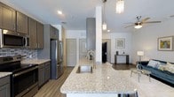 This single-floor residence showcases a thoughtfully designed open-concept layout that seamlessly connects the kitchen and living areas. The kitchen is a standout feature, anchored by a expansive granite-topped island with an undermount sink and a sleek pull-down faucet, offering generous prep and gathering space. Dark gray cabinetry provides ample storage and pairs elegantly with stainless steel appliances, including a French door refrigerator. Stylish pendant lighting with glass fixtures illuminates the island, adding a refined decorative touch.

The open flow leads naturally into the living area, where warm-toned hardwood-style flooring runs consistently throughout, unifying the space. A ceiling fan keeps the area comfortable, while recessed lighting ensures even illumination across both zones. Neutral white walls and gray accents create a clean, contemporary palette throughout.

The layout includes multiple doorways visible from the main living space, suggesting easy access to additional rooms. A small media or console area is positioned in the living room, offering practical functionality. Overall, the space delivers a modern, well-appointed aesthetic with cohesive design elements and a comfortable, airy atmosphere.