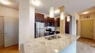 This single-floor residence offers a thoughtfully designed layout that balances modern finishes with functional living spaces. The kitchen serves as a central highlight, featuring rich dark-stained cabinetry extending to the ceiling, light granite countertops with subtle veining, and a full suite of stainless steel appliances including a refrigerator, electric range, and built-in microwave. A spacious island with an undermount double sink and elegant pendant lighting above adds both style and practicality to the space. Light hardwood-style flooring runs through the kitchen and living areas, lending warmth and continuity throughout. The living area is bright and open, benefiting from natural light that pours in through a large sliding glass door leading to an outdoor balcony. Two well-proportioned bedrooms are finished with plush neutral carpeting and windows that invite natural light. The unit includes two bathrooms: one featuring a walk-in glass-enclosed shower, granite vanity countertop, and dark wood cabinetry, and a second with a full soaking tub and shower combination, matching granite counters, and tile flooring. Clean, neutral wall tones and consistent trim details throughout create a cohesive and move-in ready aesthetic.
