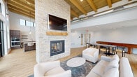 This thoughtfully designed single-floor communal space blends modern industrial and warm organic elements throughout. The main gathering area commands immediate attention with its soaring ceiling, where exposed wooden beams and visible ductwork create a distinctive architectural character. A floor-to-ceiling stacked stone fireplace serves as the room's focal point, topped with a wooden mantel and a wall-mounted television, and flanked by a comfortable seating arrangement of contemporary upholstered chairs and sofas arranged over area rugs on light hardwood flooring.

Expansive black-framed windows along multiple walls flood the interior with abundant natural light and provide open views of the surrounding outdoor area, including a patio with exterior seating. The kitchen area features rich dark wood cabinetry, open floating shelves, a textured tile backsplash, stainless steel appliances including a refrigerator and dishwasher, and a generous island beneath a trio of statement pendant lights.

A dedicated games room offers a full-length shuffleboard table alongside seating and direct access to the exterior through a glass-paneled door. A dining area with a dark wood table completes the social spaces, making the overall layout well-suited for gathering, entertaining, and relaxation.