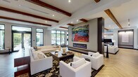 This expansive single-floor amenity space blends contemporary design with warm architectural details to create a welcoming and upscale environment. The interior is immediately distinguished by striking exposed wood ceiling beams that contrast beautifully against bright white ceilings, complemented by recessed lighting throughout. Herringbone-patterned wood flooring flows seamlessly across the main gathering areas, while large-format tile anchors the entry and transitional zones.

The grand entry lobby features a statement chandelier, floor-to-ceiling glass doors, and patterned area rugs that set an inviting tone. Multiple lounge areas are thoughtfully arranged throughout the space, each with its own character — from plush white sectionals centered around a sleek linear fireplace adorned with bold abstract artwork, to a cozy leather seating area paired with a built-in bookshelf and eclectic wall art. A dedicated media lounge with a wall-mounted television provides an additional entertainment-focused gathering spot.

Glass-enclosed private rooms with sliding barn doors offer flexible, semi-private spaces. A well-appointed full kitchen features dark cabinetry, white countertops, and stainless steel appliances. A separate coffee and refreshment station with dark lower cabinets and colorful artwork serves as a convenient amenity. Large windows throughout the space invite abundant natural light and views of lush outdoor surroundings.