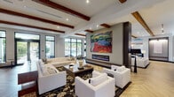 This expansive single-floor amenity space blends contemporary design with warm architectural details to create a welcoming and upscale environment. The interior is immediately distinguished by striking exposed wood ceiling beams that contrast beautifully against bright white ceilings, complemented by recessed lighting throughout. Herringbone-patterned wood flooring flows seamlessly across the main gathering areas, while large-format tile anchors the entry and transitional zones.

The grand entry lobby features a statement chandelier, floor-to-ceiling glass doors, and patterned area rugs that set an inviting tone. Multiple lounge areas are thoughtfully arranged throughout the space, each with its own character — from plush white sectionals centered around a sleek linear fireplace adorned with bold abstract artwork, to a cozy leather seating area paired with a built-in bookshelf and eclectic wall art. A dedicated media lounge with a wall-mounted television provides an additional entertainment-focused gathering spot.

Glass-enclosed private rooms with sliding barn doors offer flexible, semi-private spaces. A well-appointed full kitchen features dark cabinetry, white countertops, and stainless steel appliances. A separate coffee and refreshment station with dark lower cabinets and colorful artwork serves as a convenient amenity. Large windows throughout the space invite abundant natural light and views of lush outdoor surroundings.