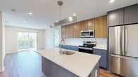 This single-floor residence showcases a thoughtfully designed contemporary interior with a cohesive aesthetic throughout. The kitchen serves as a focal point of the space, featuring a large center island with a white quartz countertop, an undermount sink, and a sleek pendant light fixture overhead. Two-toned cabinetry combines warm wood upper cabinets with slate-blue lower units, complemented by a white subway tile backsplash and a full suite of stainless steel appliances, including a refrigerator, electric range, microwave, and dishwasher. Warm-toned hardwood flooring extends seamlessly from the kitchen through the living area, which benefits from abundant natural light and direct access to a private balcony through sliding glass doors, offering outdoor views of the surrounding neighborhood. The bedroom includes a dedicated closet with built-in racking and connects to additional rooms via a bright hallway. The bathroom presents a clean, minimalist design with light wood-look tile flooring, a floating dark vanity with an integrated sink, a large mirror, and a full soaker tub with shower. Recessed lighting and white walls throughout contribute to a bright, airy ambiance.