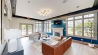 This clubhouse in Fort Wayne, Indiana, offers a thoughtfully designed gathering space that blends comfort with contemporary style. The main living area is the centerpiece of the space, featuring light hardwood flooring, bold teal accent walls, and dark crown molding that frames the high ceilings with striking contrast. A white-mantled fireplace anchors one wall, while a wall-mounted flat-screen TV adds a modern touch. Generous windows flood the room with natural light and provide pleasant outdoor views of the surrounding greenery. Seating options include a rich cognac leather sofa, complementary accent chairs, and a cozy corner nook with a marble-topped table — ideal for casual conversation or informal meetings. A large ornate mirror and a dark sideboard with gold hardware add refined decorative touches. The kitchen is fully equipped with stainless steel appliances, including a refrigerator, range, and microwave, alongside dark lower cabinetry, white upper cabinets, subway tile backsplash, and a spacious island with a deep undermount sink illuminated by matte black pendant lights. Throughout the space, curated artwork and tasteful accessories contribute to an inviting, upscale ambiance that serves as a welcoming hub for the community.