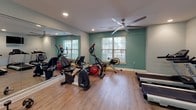 This fitness center is a well-appointed, single-floor facility designed to support a comprehensive workout experience. The space is finished with durable wood-look flooring throughout, neutral gray and teal accent walls, and recessed lighting complemented by ceiling fans that maintain comfortable airflow during exercise.

The cardio zone is anchored by treadmills, elliptical trainers, and stationary bikes, all positioned near windows dressed with slatted blinds that allow natural light to filter into the space. A large wall-to-wall mirror in this area enhances the sense of openness while allowing users to monitor their form.

The strength training section features a multi-functional cable and weight machine alongside a leg press unit, accommodating a variety of resistance exercises. A dumbbell rack provides free weight options, rounding out the strength equipment available. A mounted flat-screen television is positioned conveniently for viewing during workouts.

Additional amenities include a water fountain, a dedicated storage area, and clear exit access. The overall layout is open and organized, offering ample space to move between equipment comfortably. The combination of natural light, ceiling fans, and thoughtful equipment placement creates a functional and welcoming environment for fitness activity.