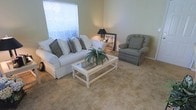 This single-floor residence offers a calm and inviting atmosphere, anchored by a thoughtfully arranged living area. The living room is finished with wall-to-wall carpeting in a warm neutral tone, contributing to a soft and comfortable underfoot feel throughout the space. Walls are painted in a light sage green, lending the room a serene, airy quality that pairs naturally with the understated décor.

A large window fitted with horizontal blinds allows natural light to filter into the living area, brightening the space while maintaining privacy. The room is spacious enough to accommodate a full-size slipcovered sofa accented with decorative pillows, a cushioned armchair, and a white glass-top coffee table — all arranged to encourage easy conversation and relaxed living.

A side table with a classic lamp and framed artwork adds a finishing touch to the room's cohesive, understated aesthetic. The overall design of the space prioritizes comfort and functionality, with clean lines and a muted color palette that creates a tranquil residential environment. The single-floor layout offers ease of accessibility and flow, making this a practical and pleasant space to occupy.