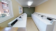 This shared laundry facility is a thoughtfully designed, single-floor space that accommodates a generous number of top-load washing machines and dryers arranged in parallel rows along both walls, maximizing capacity and ease of use. The room features a clean, neutral color palette with warm beige walls complemented by a bold deep green accent wall, giving the space a polished, intentional aesthetic. Crown molding runs along the ceiling perimeter, adding a refined architectural detail that elevates the functional nature of the room. Overhead flush-mount lighting ensures the space is well-lit throughout, while multiple windows along the walls allow natural light to filter in, offering views of the surrounding exterior. The floor is finished with a durable speckled vinyl tile, well-suited for a high-traffic utility environment. A built-in laminate folding counter runs along one wall, providing a convenient surface for sorting and folding laundry. The machines are modern and uniform in appearance, reflecting a well-maintained facility. A six-panel white door provides access to an adjacent area, which appears to connect to a mailbox alcove, suggesting this laundry room is part of a larger shared amenity space within a residential building. The overall ambiance is clean, functional, and well-organized.