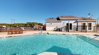 This outdoor pool area is designed with both relaxation and functionality in mind, offering a well-appointed retreat for residents. The centerpiece is a generously sized swimming pool with crystal-clear blue water, complemented by a smaller wading or spa pool nearby. The surrounding concrete deck provides ample space for lounging and sunbathing under open skies.

A covered pavilion anchors the space architecturally, featuring a warm wood-paneled ceiling, ceiling fans, and substantial natural stone columns that add a refined, durable character to the outdoor environment. The tile-covered floor of the pavilion creates a polished transition between the interior amenity building and the pool deck. Seating is available both within the shaded pavilion and along the exterior walls of the clubhouse-style building, which features a combination of board-and-batten siding and natural stone base detailing.

Beyond the pool, a gravel-lined area extends toward a wrought iron perimeter fence, with an outdoor dining set positioned beneath a large market umbrella offering views of mature trees and open sky. The overall ambiance is clean, modern, and inviting, balancing durable outdoor materials with thoughtful design details suited for year-round enjoyment.
