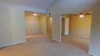 This single-floor residence is designed with an open and airy feel, highlighted by striking vaulted ceilings that lend a sense of generous vertical space to the main living area. The interior is finished with warm, neutral-toned walls that create a welcoming and cohesive atmosphere throughout. Soft, plush wall-to-wall carpeting spans the living spaces, adding comfort underfoot and contributing to the home's understated, cozy character. The open-concept layout connects the main living area seamlessly to adjoining spaces, allowing for flexible use and easy flow between rooms. Recessed lighting provides even, ambient illumination, complementing the natural brightness of the space. A dedicated area visible from the main room appears to function as an additional living or flex space, offering versatility for various uses. A hallway leads to what appears to be a bedroom with a closet, indicated by a bifold or panel door — a practical and well-integrated storage solution. HVAC vents are visibly positioned to ensure climate comfort throughout the unit. The overall design emphasizes clean lines, neutral finishes, and functional simplicity, making this a move-in-ready space well-suited for a variety of lifestyles.