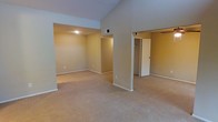 This single-floor residence is designed with an open and airy feel, highlighted by striking vaulted ceilings that lend a sense of generous vertical space to the main living area. The interior is finished with warm, neutral-toned walls that create a welcoming and cohesive atmosphere throughout. Soft, plush wall-to-wall carpeting spans the living spaces, adding comfort underfoot and contributing to the home's understated, cozy character. The open-concept layout connects the main living area seamlessly to adjoining spaces, allowing for flexible use and easy flow between rooms. Recessed lighting provides even, ambient illumination, complementing the natural brightness of the space. A dedicated area visible from the main room appears to function as an additional living or flex space, offering versatility for various uses. A hallway leads to what appears to be a bedroom with a closet, indicated by a bifold or panel door — a practical and well-integrated storage solution. HVAC vents are visibly positioned to ensure climate comfort throughout the unit. The overall design emphasizes clean lines, neutral finishes, and functional simplicity, making this a move-in-ready space well-suited for a variety of lifestyles.