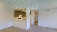 This single-story space is designed with a practical, open layout that connects the kitchen and living areas seamlessly. The kitchen is equipped with medium-toned wooden cabinetry that provides ample storage, a gas range, and a white refrigerator. A peninsula-style countertop with a raised bar section separates the kitchen from the living area, creating a natural division while maintaining an open feel. The living area features neutral wall-to-wall carpeting in a warm beige tone that extends throughout the space, lending a cohesive and comfortable feel underfoot. A textured popcorn ceiling runs throughout, adding a classic residential character. Adjacent to the main living area, a hallway leads to additional rooms, suggesting a well-organized layout with clear separation between common and private spaces. The overall color palette is neutral, with white walls and warm wood accents creating a clean, understated aesthetic. Recessed lighting in the kitchen and flush-mount ceiling fixtures in the hallway provide consistent illumination throughout. The space offers a functional and straightforward design suited to everyday living.