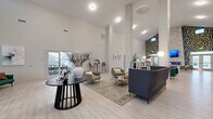 Located at 5813 Lilliana Ln in Whitestown, Indiana, this single-floor community clubhouse offers a generously sized, open-plan gathering space designed with modern aesthetics and functional comfort in mind. The interior immediately impresses with its soaring double-height ceilings, which lend an airy, expansive feel throughout the space. Light wood-style flooring runs continuously underfoot, complementing the bright, neutral color palette of the walls and architectural elements.

The central lounge area serves as the heart of the space, anchored by a bold, slatted round table and surrounded by carefully arranged seating groupings — including plush sofas, accent chairs, and area rugs — ideal for relaxed socializing or informal gatherings. A striking accent wall features a combination of stacked stone and bold geometric patterned tile, reaching the full height of the room and serving as a dramatic focal point alongside French doors that invite natural light from outside.

A fully equipped kitchen and bar area offers white upper cabinetry, slate-gray lower cabinets, stainless steel appliances, a large quartz-topped island with bar seating, pendant lighting, and a beverage station — well-suited for events and everyday resident use. Additional seating nooks and entry vestibules with large glass doors and windows further enhance the bright, welcoming atmosphere of this thoughtfully designed community amenity space.
