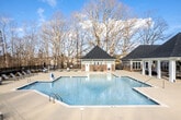 Located at 114 Avalon Park Rd in Mooresville, North Carolina, this single-story community space offers a thoughtfully designed environment that blends classic architectural style with comfortable outdoor living. The exterior showcases a timeless combination of brick masonry and sage-green lap siding, accented by crisp white trim and traditional rooflines that create a warm, residential character.

A generously sized covered patio serves as a central gathering area, supported by sturdy white columns and equipped with ceiling fans to ensure comfort year-round. The patio is furnished with wicker seating, including cushioned chairs and lounge ottomans, making it an inviting space for relaxation and socializing. Double glass-paned entry doors with classic wall lanterns provide an elegant focal point to the building entrance.

The outdoor area extends to a expansive resort-style swimming pool with a broad aggregate concrete deck, lounge chairs, and umbrella-shaded seating areas. The pool is bordered by decorative metal fencing, with views of surrounding mature trees that provide a natural, serene backdrop.

Additional building structures on the property mirror the same brick-and-siding aesthetic, connected by wide, open concrete walkways that facilitate easy movement throughout the campus. The overall ambiance is one of a well-appointed, community-oriented environment designed for comfort, accessibility, and outdoor enjoyment.