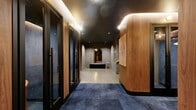 Located on the 23rd floor of the iconic 95 Wall Street in Lower Manhattan, this 720-square-foot co-working suite delivers a polished and professional environment designed for productivity. The space encompasses a hallway, a primary meeting room, a bathroom, and two additional rooms suited for focused work or collaboration.

The hallway sets an immediate tone of sophistication, featuring warm walnut wood-paneled walls, a dark coffered ceiling with soft cove lighting, and a striking blue-toned carpet floor offset by large-format stone tile. Curated artwork, including a bold abstract canvas and a vintage typewriter print, adds character to the corridor.

Flanking the hallway are sleek private phone booths enclosed in black-framed glass doors with ribbed glazing panels, providing acoustic privacy for calls or focused work. Each booth is outfitted with seating, creating intimate, self-contained workspaces.

The primary meeting room features a round stone-topped table surrounded by plush blush-pink curved armchairs, set against deep charcoal walls. Ribbed glass partitions allow light to filter through while maintaining a sense of enclosure. The overall ambiance throughout the suite is contemporary and refined, balancing dark, moody tones with warm wood accents and carefully layered lighting to create an upscale, focused workspace in the heart of New York City's Financial District.