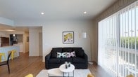 This single-floor space presents a thoughtfully designed living area that balances modern aesthetics with functional comfort. The living room serves as the focal point, featuring a dark upholstered sofa accented with vibrant, patterned throw pillows that add a playful contrast to the neutral color palette. A round white coffee table anchors the seating area, complemented by a plush white area rug that defines the space. A bold pop-art framed print adds a striking visual element to the clean white walls, lending the room a contemporary and artistic character. Recessed ceiling lights provide even, ambient illumination throughout, while a sleek arc floor lamp contributes additional warm lighting near the seating area. Large windows dressed with sheer vertical blinds allow natural light to filter generously into the room, creating a bright and welcoming atmosphere. The light wood-tone flooring runs throughout, adding warmth and continuity to the space. An open sightline toward an adjoining hallway suggests a practical and well-connected layout. Overall, the space conveys a clean, modern sensibility with curated décor choices.