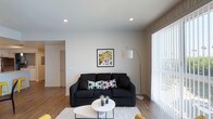 This single-floor space presents a thoughtfully designed living area that balances modern aesthetics with functional comfort. The living room serves as the focal point, featuring a dark upholstered sofa accented with vibrant, patterned throw pillows that add a playful contrast to the neutral color palette. A round white coffee table anchors the seating area, complemented by a plush white area rug that defines the space. A bold pop-art framed print adds a striking visual element to the clean white walls, lending the room a contemporary and artistic character. Recessed ceiling lights provide even, ambient illumination throughout, while a sleek arc floor lamp contributes additional warm lighting near the seating area. Large windows dressed with sheer vertical blinds allow natural light to filter generously into the room, creating a bright and welcoming atmosphere. The light wood-tone flooring runs throughout, adding warmth and continuity to the space. An open sightline toward an adjoining hallway suggests a practical and well-connected layout. Overall, the space conveys a clean, modern sensibility with curated décor choices.