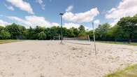Located at 1715 Lake Michigan Drive in Valparaiso, Indiana, this outdoor recreational amenity area offers a well-rounded selection of sports and leisure facilities set against a backdrop of lush, mature trees and manicured green space. The grounds feature multiple sand volleyball courts equipped with regulation nets and sturdy pole anchors, providing an ideal surface for casual and competitive play alike. Adjacent to the volleyball area, a full basketball court with a mounted backboard and hoop offers a paved surface complete with painted court markings, accommodating both half-court and full-court play. Tennis courts enclosed by chain-link fencing provide a dedicated space for racket sports, with clearly marked lines and surrounding greenery adding to the open-air atmosphere. Open lawn areas with decorative boulders and neatly spaced trees contribute to the overall parklike setting. Paved pathways connect the various amenity zones, and lighting poles suggest availability for evening use. The entire complex is framed by views of the surrounding residential community, with a bright, open sky overhead enhancing the welcoming, active outdoor environment throughout all seasons.