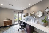 Located at 1239 Bay Area Blvd in Houston, Texas, this single-room business center is a thoughtfully designed workspace that balances functionality with contemporary style. The room is anchored by a long, continuous marble-topped desk that spans nearly the full length of one wall, accommodating multiple workstations equipped with desktop computers and ergonomic task chairs. Recessed lighting overhead ensures consistent, well-distributed illumination throughout the space. A striking decorative feature on the accent wall showcases an eclectic collection of wall-mounted clocks alongside industrial-style pipe shelving, which displays greenery, framed certifications, and curated décor items — lending the room a distinctive character. Natural light flows in generously through floor-to-ceiling double glass doors fitted with roller shades, offering views of a landscaped outdoor area with mature trees and manicured greenery. A rustic wood-and-white storage chest positioned near the doors adds a warm, textural contrast to the room's modern grey palette. The floor is finished with a vibrant geometric patterned carpet in shades of blue, yellow, and grey, adding an energetic visual element underfoot. An open entryway connects the business center to an adjacent common area, reinforcing its role as part of a larger amenity offering within this Houston property.