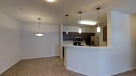This single-floor residence is designed with an open-concept layout that maximizes the sense of space and connectivity between the kitchen and living areas. The kitchen is a standout feature, equipped with stainless steel appliances including a refrigerator, microwave, and range. Rich, dark espresso-toned cabinetry lines the walls, offering ample storage, while the crisp white peninsula counter provides additional workspace and doubles as a casual dining or entertaining surface. Three stylish pendant lights hang above the peninsula, casting a warm, focused glow over the area and adding a contemporary decorative touch. A coordinating bowl-style pendant light illuminates the adjacent living space, maintaining a cohesive aesthetic throughout. The flooring throughout the space is a warm hardwood-style material that adds texture and continuity, tying each area together seamlessly. The neutral wall tones and clean architectural lines create a bright, welcoming ambiance. The open flow between the kitchen and living zone makes the layout highly functional, ideal for both everyday living and entertaining guests.