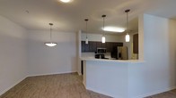 This single-floor residence is designed with an open-concept layout that maximizes the sense of space and connectivity between the kitchen and living areas. The kitchen is a standout feature, equipped with stainless steel appliances including a refrigerator, microwave, and range. Rich, dark espresso-toned cabinetry lines the walls, offering ample storage, while the crisp white peninsula counter provides additional workspace and doubles as a casual dining or entertaining surface. Three stylish pendant lights hang above the peninsula, casting a warm, focused glow over the area and adding a contemporary decorative touch. A coordinating bowl-style pendant light illuminates the adjacent living space, maintaining a cohesive aesthetic throughout. The flooring throughout the space is a warm hardwood-style material that adds texture and continuity, tying each area together seamlessly. The neutral wall tones and clean architectural lines create a bright, welcoming ambiance. The open flow between the kitchen and living zone makes the layout highly functional, ideal for both everyday living and entertaining guests.