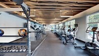 Located at 1225 Willowbrook Dr SE in Huntsville, Alabama, this single-floor fitness center offers a generously sized, open workout environment designed to accommodate a wide range of exercise needs. The space is defined by its striking exposed wood-slat ceiling, which adds a warm, industrial-chic aesthetic, complemented by exposed ductwork and pendant lighting that illuminates the floor below.

The facility is divided into distinct workout zones. Along one side, a long row of cardio equipment — including treadmills, stationary bikes, ellipticals, and stair climbers — lines the large windows that face the outdoors, providing natural light and pleasant exterior views during workouts. On the opposite end, a dedicated strength training area houses power racks, cable machines, a full dumbbell rack, kettlebells, battle ropes, a punching bag, and various selectorized weight machines. Wall-mounted TVs are positioned throughout for entertainment.

A separate, open floor zone with rubber flooring provides space for functional training and stretching, stocked with medicine balls, stability balls, and adjustable benches. Glass-panel doors connect the gym to an adjacent area, adding a sense of openness. The light gray walls, rubber flooring throughout, and abundant natural light from large windows give the space a clean, modern, and energizing atmosphere.