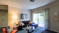 This single-floor residence features a thoughtfully designed living room that serves as the centerpiece of the space. The room is finished with smooth, light-toned walls accented by elegant crown molding, lending a polished, refined character to the interior. A large sliding glass door dominates one wall, allowing abundant natural light to fill the space while providing direct access to an outdoor patio area, where glimpses of greenery and stone detailing are visible beyond.

The living room is styled with a dark sectional sofa paired with an assortment of patterned throw pillows in warm orange and neutral tones, complemented by a sleek glass-top coffee table and a geometric-patterned area rug. Patterned curtains frame the sliding door, adding visual texture and softness. A colorful abstract canvas artwork adorns the wall, introducing vibrant blue, orange, and white tones to the neutral palette. A floor lamp positioned in the corner contributes warm ambient lighting.

Ceiling-mounted HVAC ventilation is centrally located above, ensuring consistent climate control throughout. The overall ambiance is modern, airy, and inviting, blending functional design with contemporary décor sensibilities.