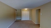 This single-floor unit presents a straightforward and functional layout designed for comfortable everyday living. The main living area is finished with wall-to-wall carpeting in a warm neutral tone, providing a soft underfoot feel throughout the space. Walls are painted in a muted beige hue, contributing to a calm and understated ambiance that serves as a versatile backdrop for a variety of interior styles. The room features a textured ceiling with a centrally placed flush-mount light fixture that provides general illumination to the space. A set of bifold closet doors offers built-in storage, neatly integrated into the wall to maximize usable floor space. A solid entry door with standard hardware provides access to the unit, framed by clean white trim consistent with the room's overall aesthetic. An open passage on one side suggests a connected area or additional room, hinting at a practical floor plan designed to make efficient use of the available space. White baseboards run along the perimeter, adding a crisp finishing detail that ties together the neutral color palette. Overall, the space is clean, move-in ready, and offers a blank canvas well-suited for personalization.