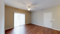 This 1-bedroom, 1-bathroom residence is thoughtfully laid out across a single floor, offering a comfortable and functional living environment. The living area is a standout feature, showcasing rich, dark hardwood-style flooring that adds warmth and elegance throughout the space. A ceiling fan with an integrated light fixture is centrally positioned, providing both air circulation and ambient lighting. A large sliding glass door floods the room with natural light and opens to an outdoor view, creating a bright and airy atmosphere. Neutral-toned walls in a soft beige complement the flooring and contribute to the space's clean, welcoming aesthetic. White baseboards and trim provide a crisp contrast throughout. A closet door is visible, indicating convenient storage access within the living area. The overall design balances practicality with comfort, making it an ideal space for those seeking a straightforward, move-in-ready home. The single-floor plan ensures easy navigation, while the natural light and open feel of the main living area make the space feel more expansive than its footprint might suggest.