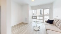 This thoughtfully designed 1-bedroom, 2-bathroom unit delivers a clean, modern aesthetic across a single floor. Light-toned hardwood flooring runs throughout, tying the spaces together seamlessly. The living room is a bright, airy retreat anchored by expansive floor-to-ceiling windows and a sliding glass door that opens onto a private balcony, offering open views of the surrounding urban skyline and filling the space with generous natural light.

The bedroom is a comfortable, well-proportioned room featuring its own large window with city views, a built-in closet, and the same light hardwood flooring. The two bathrooms are finished to a high standard, showcasing elegant marble-look tile on both the floors and shower/tub surrounds, paired with chrome fixtures for a polished, contemporary feel.

A hallway connects the living areas to the private quarters, and conveniently incorporates a dedicated in-unit laundry nook with a stacked washer and dryer. The kitchen, visible in the dollhouse view, features white cabinetry and stainless steel appliances. Throughout the unit, crisp white walls, recessed lighting, and clean lines create a cohesive, modern ambiance that is both functional and refined.