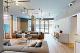 Located at 77 Bruton Street in San Francisco, this single-floor space presents a refined, contemporary living room that masterfully balances modern design with warm, natural materials. The open-plan layout maximizes the available floor area, creating a fluid and inviting atmosphere throughout.

The living room is the heart of the space, anchored by generous sectional and modular sofas in muted tones of gray and taupe that complement the room's sophisticated palette. Light hardwood flooring runs continuously underfoot, lending warmth to the otherwise cool, muted color scheme. The walls feature a distinctive layered treatment, combining smooth white lower panels with warm light wood cladding and textured concrete-effect upper sections in soft sage and gray tones — a design detail that adds depth and architectural interest.

Overhead, a dramatic arrangement of sculptural pendant and track lighting fixtures creates a striking focal point, casting ambient and accent light throughout the space. Floor-to-ceiling glass doors flood the room with natural light and provide direct access to an outdoor area, enhancing the sense of openness. A sleek built-in credenza with white cabinetry and bar seating anchors one side of the room, adding functional elegance. The space concludes with a cozy lounge nook lined with wood-paneled walls and soft uplighting, offering an intimate retreat within the broader open-plan environment.